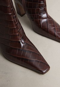 Braune knöchelhohe Stiefel aus krokodilgeprägtem Leder mit spitzer Zehenpartie, kleinem Absatz und seitlichem Reißverschluss, mit glänzender Textur.