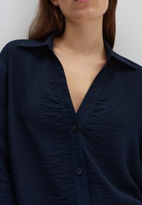 Camicia col colletto di colore blu navy con tessuto testurizzato, dotata di scollatura a V profonda, chiusura con bottoni e silhouette rilassata.