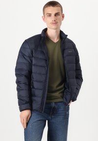 Tumši zila pūkaina jaka ar kvadrātveida rakstu, rāvējslēdzēja priekšpusi un augstu apkakli. Kombinēta ar olīvu zaļu V-veida kakla kreklu un ziliem džinsiem.