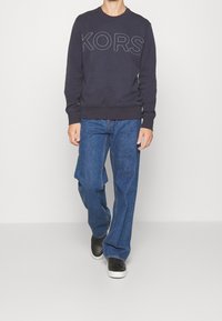 Námořnický svetr s nápisem "KORS" v kontrastním prošívání, kombinovaný s volnými modrými džíny a černými teniskami. Hedvábná tkanina s vroubkovanými manžetami.