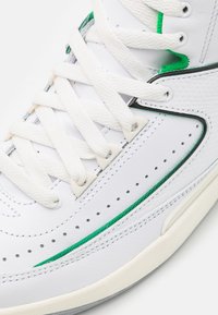 Bílé kožené tenisky se zelenými akcenty, texturovanými boky, perforovanými vzory a plochými bílými tkaničkami, s kulatou špičkou a gumovou podrážkou.