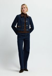 Tmavě modrá džínová bunda s hnědým límcem a kapsami, stříbrné knoflíky, v kombinaci s hnědým rolákem a rozšířenými tmavými džíny. Hnědé boty.