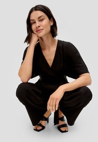 Zwarte jumpsuit met een diepe V-hals, korte mouwen en een strikceintuur. Model draagt zwarte instapsandalen, wat de gladde stof en de relaxte pasvorm laat zien.