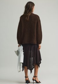 Bruine, pluizige trui, gedragen over een donkere, asymmetrische rok. Houdt een gestreept overhemd vast, draagt zwarte sandalen met bandjes. Eenvoudige, gestructureerde details.