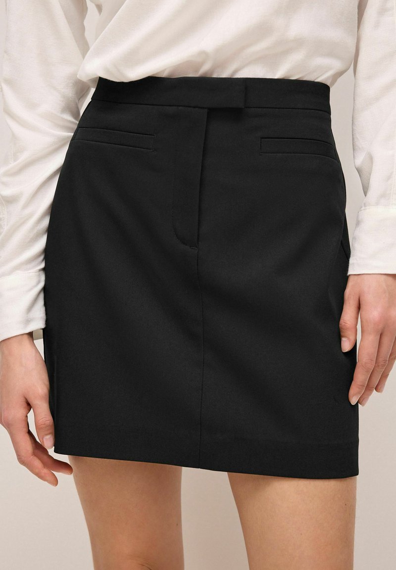 Falda negra con un diseño a medida, que presenta bolsillos frontales, una textura suave y un corte recto, combinada con una camisa blanca de manga larga.