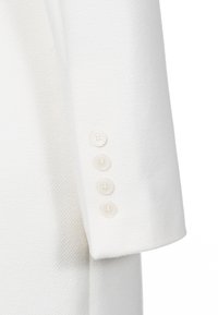 Une manche de blazer blanc présentant un tissu texturé, quatre boutons crème et un bord net et ajusté. Le design est minimaliste et élégant.