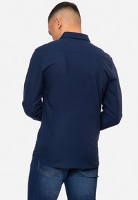 Camisa de manga larga azul marino con cuello, de textura suave y cierre de botón en los puños, vista desde la espalda. Lleva puesta unos jeans oscuros.