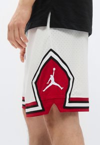 Białe siateczkowe spodenki do koszykówki z czerwonymi i czarnymi akcentami, wzorem w kształcie diamentu oraz białym logo Jumpman po lewej stronie.