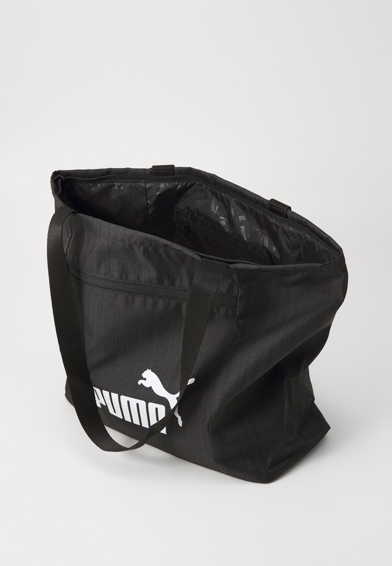Bolso de tela negro con cremallera en la parte superior, que presenta un logo blanco de Puma y resistentes correas de hombro negras. El interior tiene un forro con un diseño.