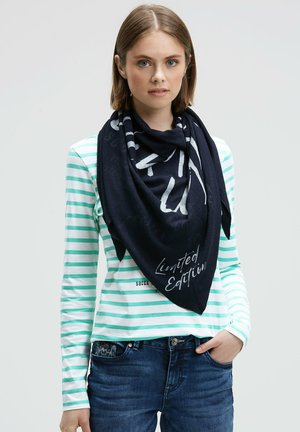 Woman with short hair wearing a navy scarf with "Limited Edition" text, a light green striped long-sleeve shirt, and dark blue jeans.
