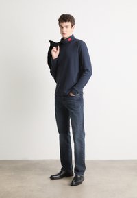Camicia a maniche lunghe blu navy con colletto alto, etichetta rossa al collo, abbinata a jeans scuri e stivali di pelle nera, in piedi su uno sfondo chiaro.