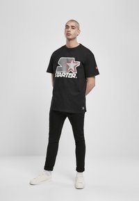 Černé tričko s grafickým logem, které obsahuje červenou hvězdu a nápis "STARTER", doplněné o černé kalhoty a bílé tenisky.