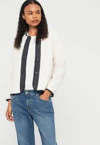 Veste blanche duveteuse avec un plastron et des finitions bleu marine, associée à un jean en denim bleu clair. Texture lisse et coupe décontractée.