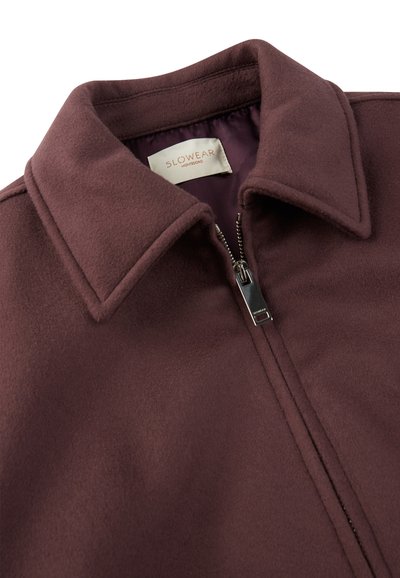 Bordeaux fleecjas met een staande kraag, voorzien van een ritssluiting en een gladde binnenvoering. Etiket zichtbaar aan de binnenkant van de kraag.
