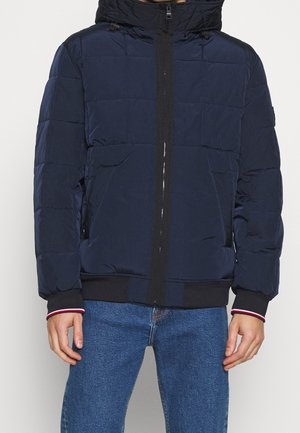 Man wearing a navy blue quilted jacket with black zipper and elastic cuffs, paired with blue jeans against a plain background.