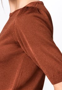 Rust-colored, textured top with three-quarter sleeves, ribbed fabric, and a subtle gathered detail at the shoulder seam.