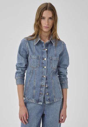 Mlada ženska, oblečena v oprijeto jeans jakno z gumbi in prsni žepki ter usklajene jeans hlače, stoji pred enobarvnim ozadjem.