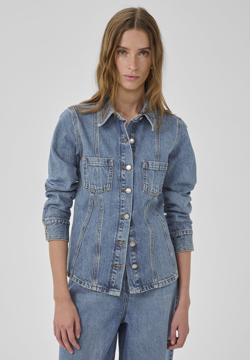 Mladá žena v priliehavej rifľovej bunde zapínanej na gombíky s náprsné vrecká a zladenými rifľovými džínsami, stojaca pred jednoliatym pozadím.