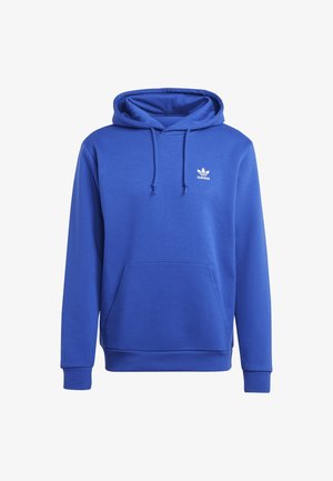 Mężczyzna ubrany w niebieską bluzę z kapturem Adidas z przednią kieszenią, czarne spodnie dresowe Adidas z białymi paskami oraz czarne buty Adidas z czerwonymi i niebieskimi akcentami.