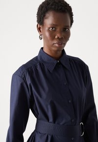 Navy blauwe shirtjurk met een gestructureerde kraag, knoopsluiting en een ceintuur in de taille. Glad materiaal met een subtiele glans en geplooide details op de schouders.