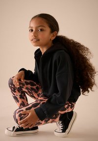 Haut à zip noir, leggings à motif animal rose et noir, baskets noires avec semelles blanches, cheveux longs et bouclés, teint naturel.