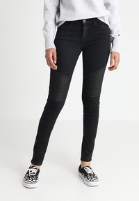 Černé skinny džíny s vroubkovanými texturovanými panely na kolenou, vyrobené z denimové látky. Nosené s vzorovanými černými teniskami.