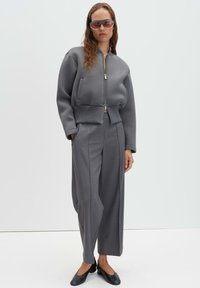 Giubbotto bomber grigio corto con chiusura a zip, abbinato a pantaloni grigi ampi e ballerine nere. Modello con occhiali da sole colorati.