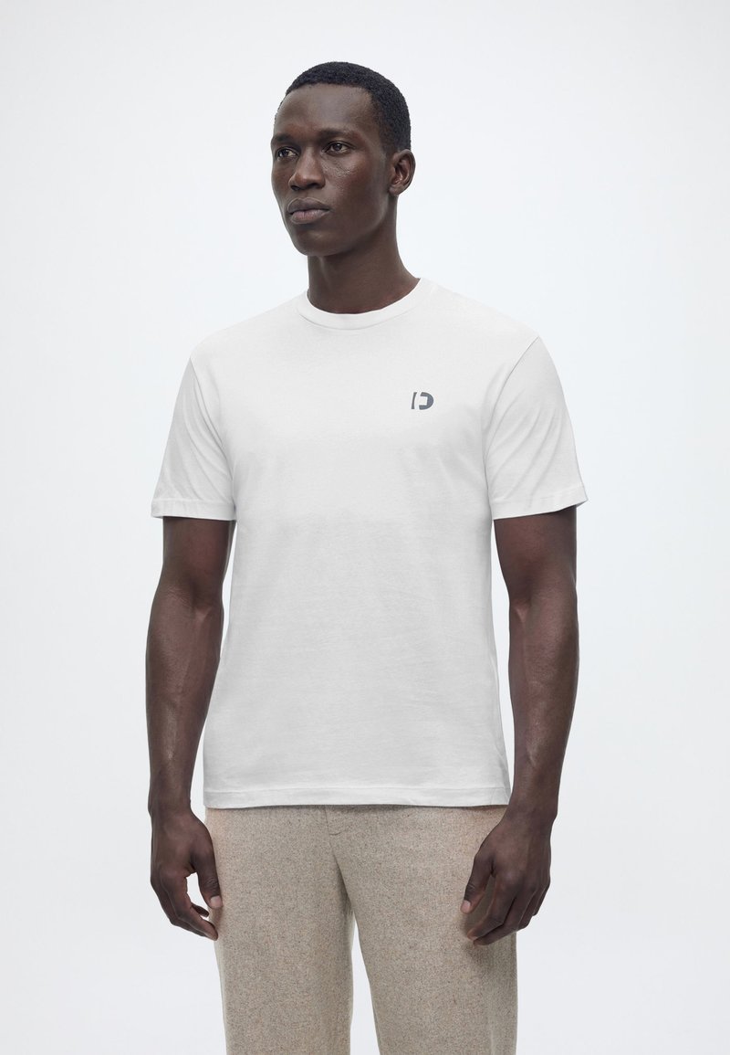 Άνδρας που φοράει απλό λευκό κοντομάνικο μπλουζάκι με μικρό γκρι λογότυπο στο αριστερό μέρος του στήθους και μπεζ παντελόνι, στέκεται μπροστά σε λευκό φόντο.