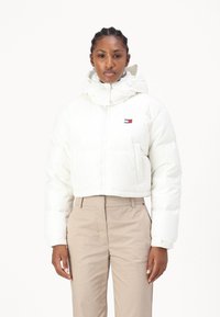 Hvit kort pufferjakke med hette, med en liten logo på brystet. Glatt materiale med kviltet tekstur; satt sammen med beige bukser.