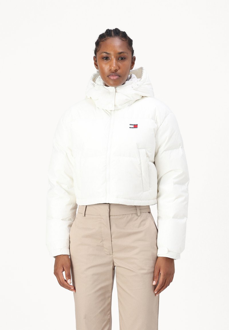 Hvit kort pufferjakke med hette, med en liten logo på brystet. Glatt materiale med kviltet tekstur; satt sammen med beige bukser.