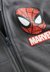 Egy szürke cipzáras ruházat közeli felvétele, amelyen egy piros és fekete Pókember arc grafika, valamint egy fehér Marvel logó található.