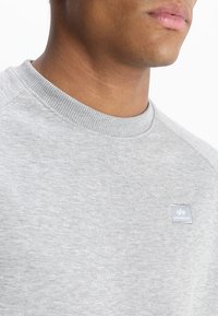 Sudadera gris con cuello y puños acanalados, que presenta una pequeña etiqueta azul con letras blancas. Textura suave y diseño clásico.