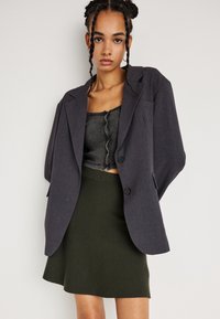 Mladá žena s vrkôčkami, oblečená v tmavom oversized saku, prúžkovanom cropped tričku s gombíkmi a olivovozelených sukni pred bielou stenou.