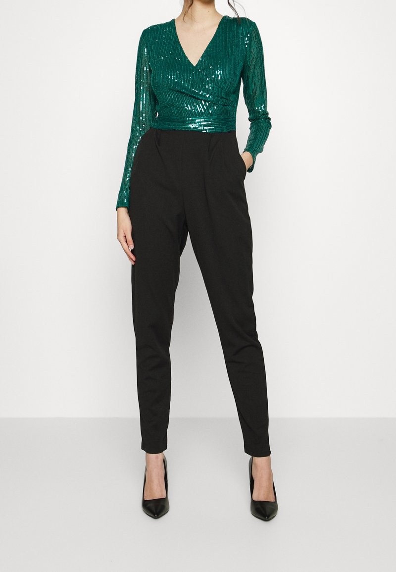Top vert à sequins avec décolleté en V et manches longues, associé à un pantalon noir ajusté et des escarpins noirs à bout pointu.