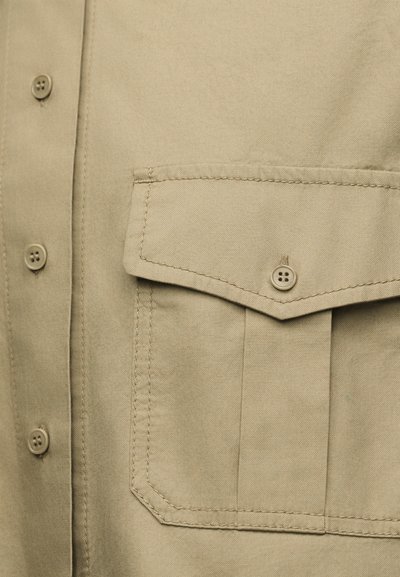 Κοντινό πλάνο σε πουκάμισο από μπεζ ύφασμα που δείχνει τσέπη με πατιλέτα και τρία κουμπιά τοποθετημένα κάθετα.