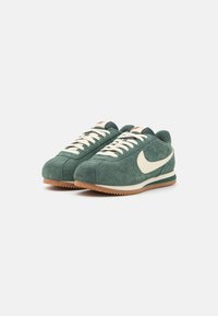 Zelené semišové tenisky s bílým logem swoosh, smetanovými tkaničkami a podrážkou z gumové pryže. Zaoblená špička a detail polstrovaného límce.