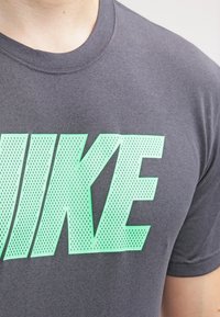 Szara koszulka z zielonym perforowanym logo "NIKE" z przodu. Materiał wydaje się lekki i gładki, z klasycznym okrągłym dekoltem.