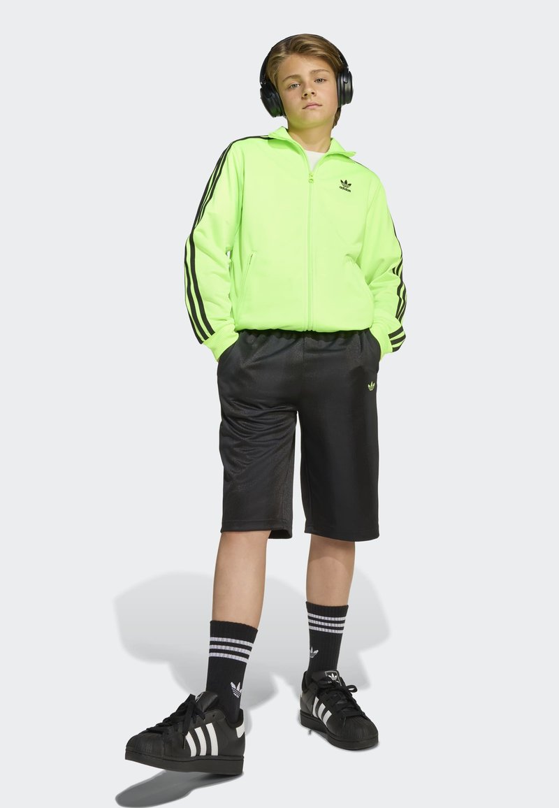 Neongrön zip-up jacka med svarta ränder, kombinerad med svarta shorts. Bärs med svarta skor, randiga strumpor och hörlurar.