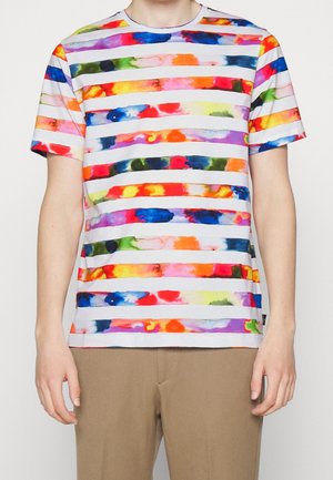 Person mit einem kurzärmeligen weißen T-Shirt mit horizontalen Streifen in bunten, aquarellähnlichen Mustern und beigen Hosen.