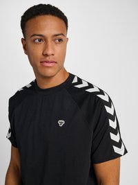 Mężczyzna z krótkimi kręconymi włosami, ubrany w czarną koszulkę z białymi pasami w kształcie szewronów na rękawach i małe logo pszczoły na piersi, patrzy w lewo.