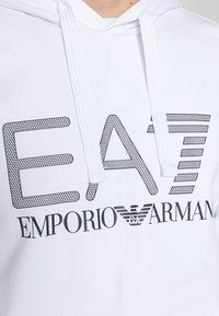 Bílá mikina s kapucí, která má velké černé logo „EA7“ se síťovaným vzorem a text „Emporio Armani“. Nastavitelné šňůrky na výstřihu. Hladká textura.