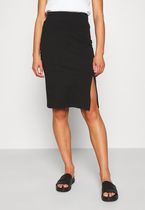 Femme portant une jupe noire longueur genou avec une fente latérale, un haut blanc et des sandales noires sans lacets, debout devant un fond blanc uni.