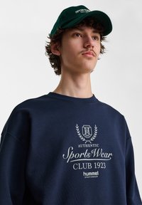 Fiatal férfi sötétzöld sapkát és tengerészkék pulóvert visel, amelyen fehér "Authentic SportsWear Club 1923" logó és felirat található.