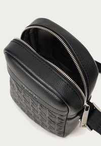 Melna ādas soma ar kvadrātveida ģeometrisko rakstu, metāla rāvējslēdzēja aizdare un regulējama siksna. Iekšpuse ir tumša un plaša.