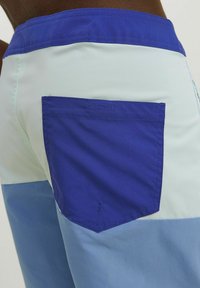 Blå och ljusgröna badshorts med en solid blå ficka på baksidan, med en slät yta och kontrasterande färgblock.