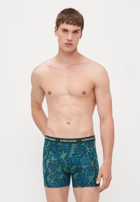Boxershorts im tropischen Blattmuster in Türkis und Dunkelgrün. Weicher Stoff mit einem schwarzen Markenbund. Mittlere Oberschenkellänge, enganliegendes Design.
