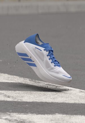 Chaussure de sport blanche et bleue suspendue au-dessus d'un passage piéton sur une route asphaltée avec de petites particules de saleté visibles en dessous.