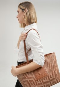 Brown tote bag with textured diagonal pattern, logo accent, and dual handles, carried by a person in a white shirt.
