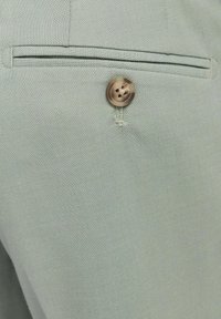 Gros plan sur un pantalon en tissu gris clair montrant un bouton marron unique au-dessus d'une poche horizontale fendue.