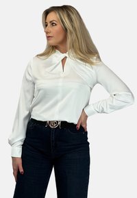 Biała bluzka z długim rękawem z detalem kołnierza w kształcie otworu, gładka tekstura i zapięcie na guziki. Połączona z ciemnymi dżinsami i paskiem z logo.